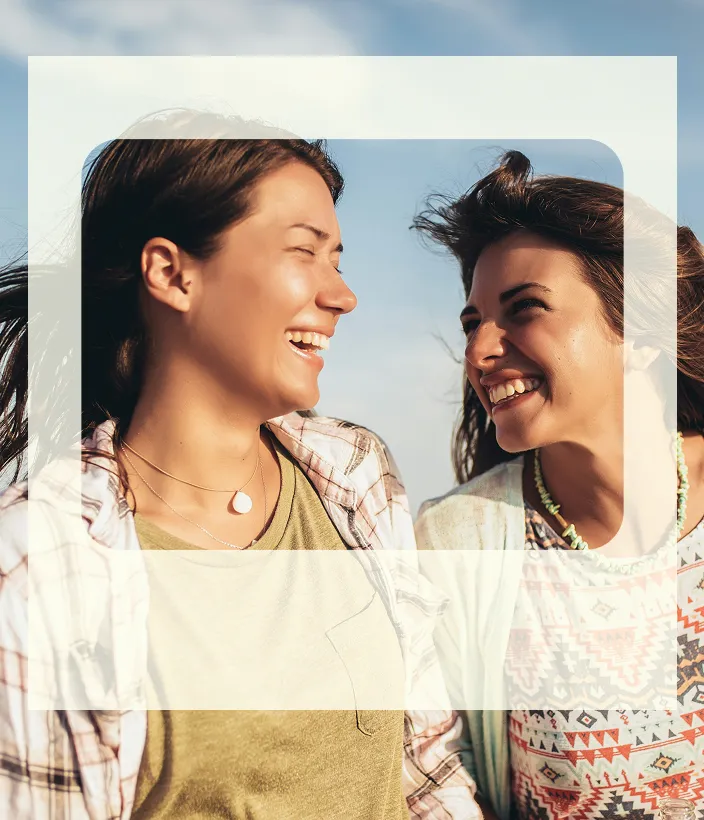 Two women smiling and laughing together outdoors with a blue sky background.