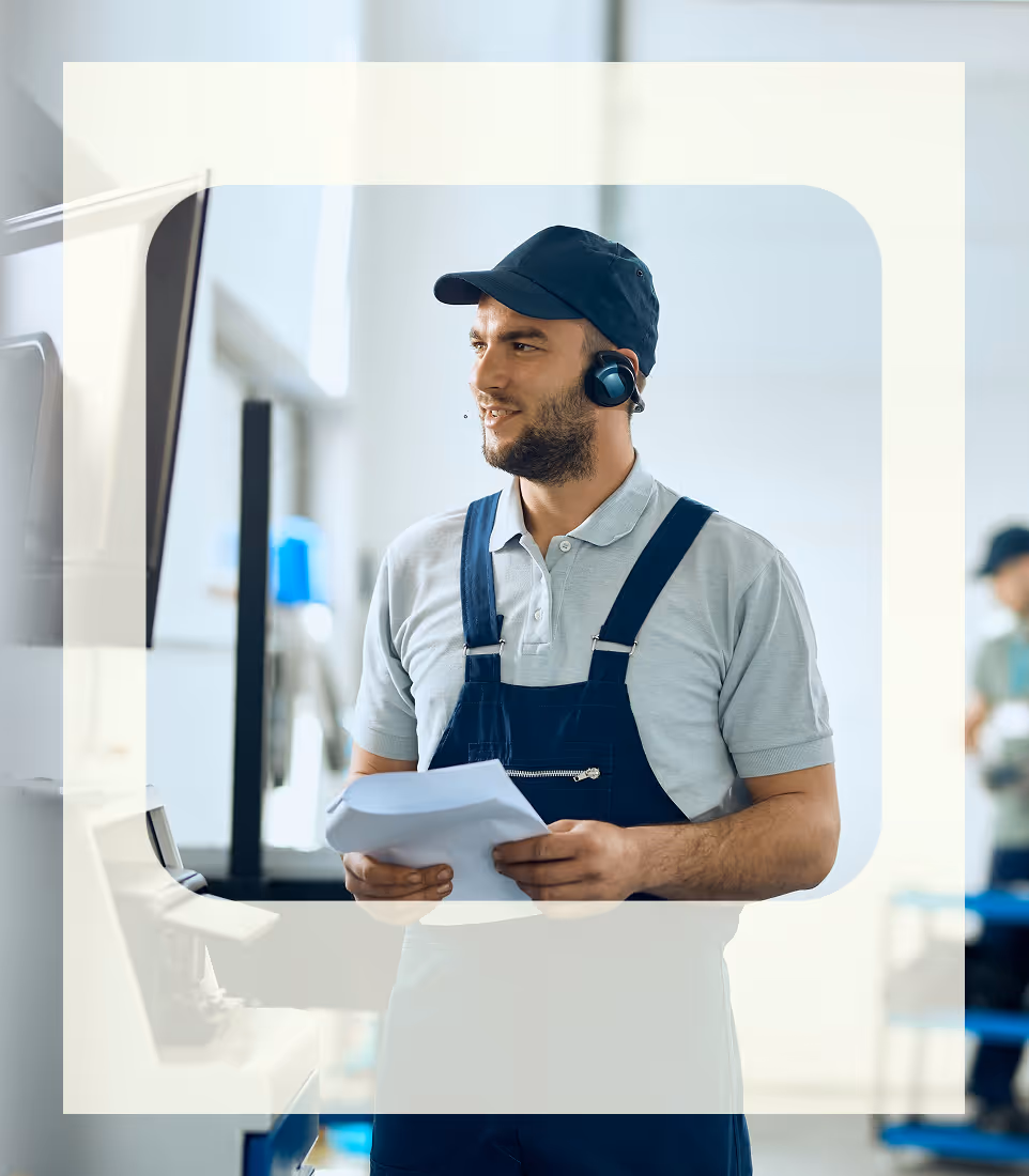 Male worker in blue overalls and cap holding papers, standing indoors near a machine with a screen.