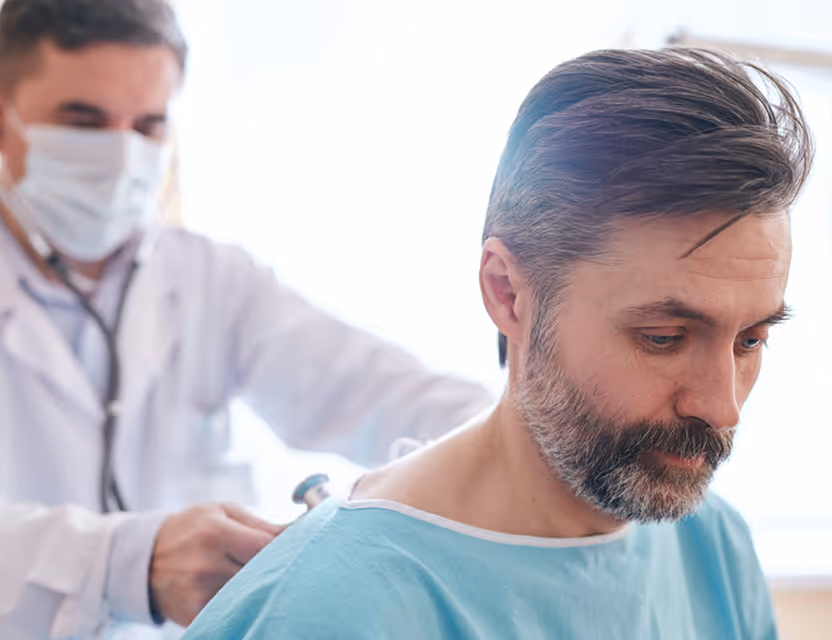 Doctor in mask using stethoscope to examine a middle-aged male patient in a medical gown.