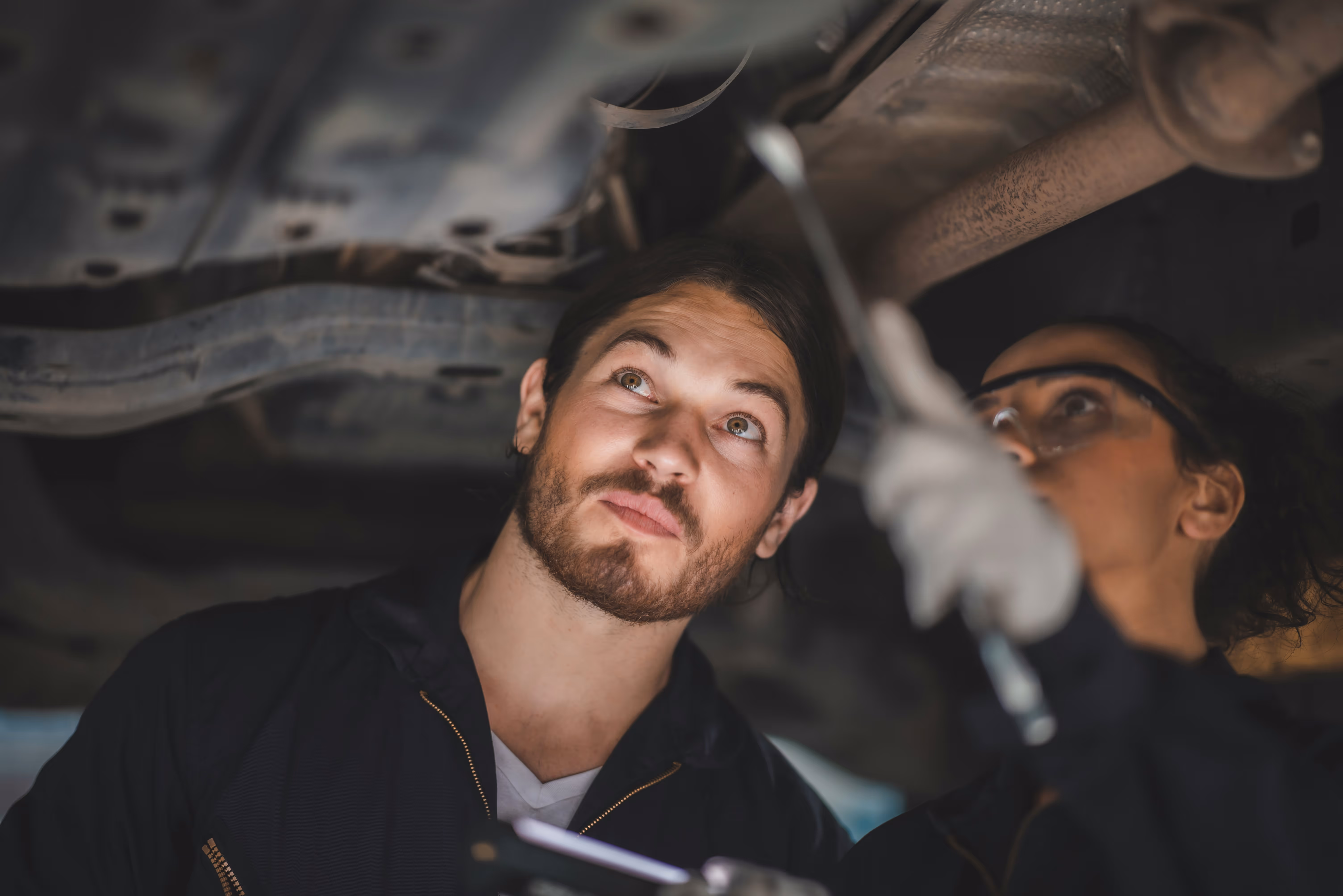 Two mechanics, one holding a wrench, inspecting the underside of a vehicle in a garage.