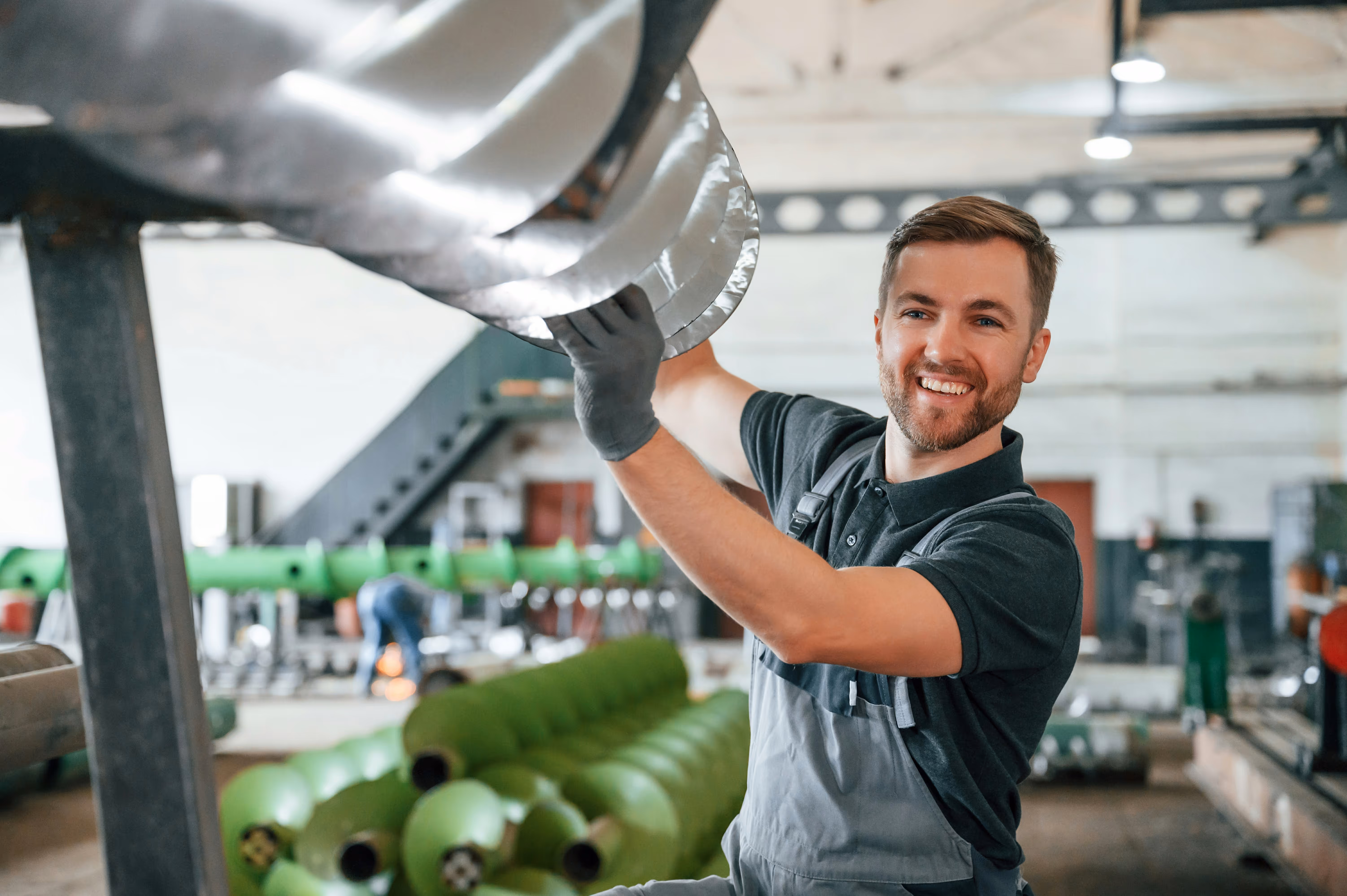 Smiling male factory worker adjusting shiny metal ducting in industrial workshop.
