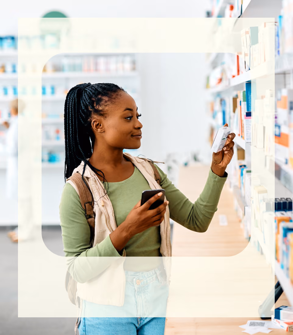 Woman in pharmacy holding a smartphone and reading a medicine box from a shelf.