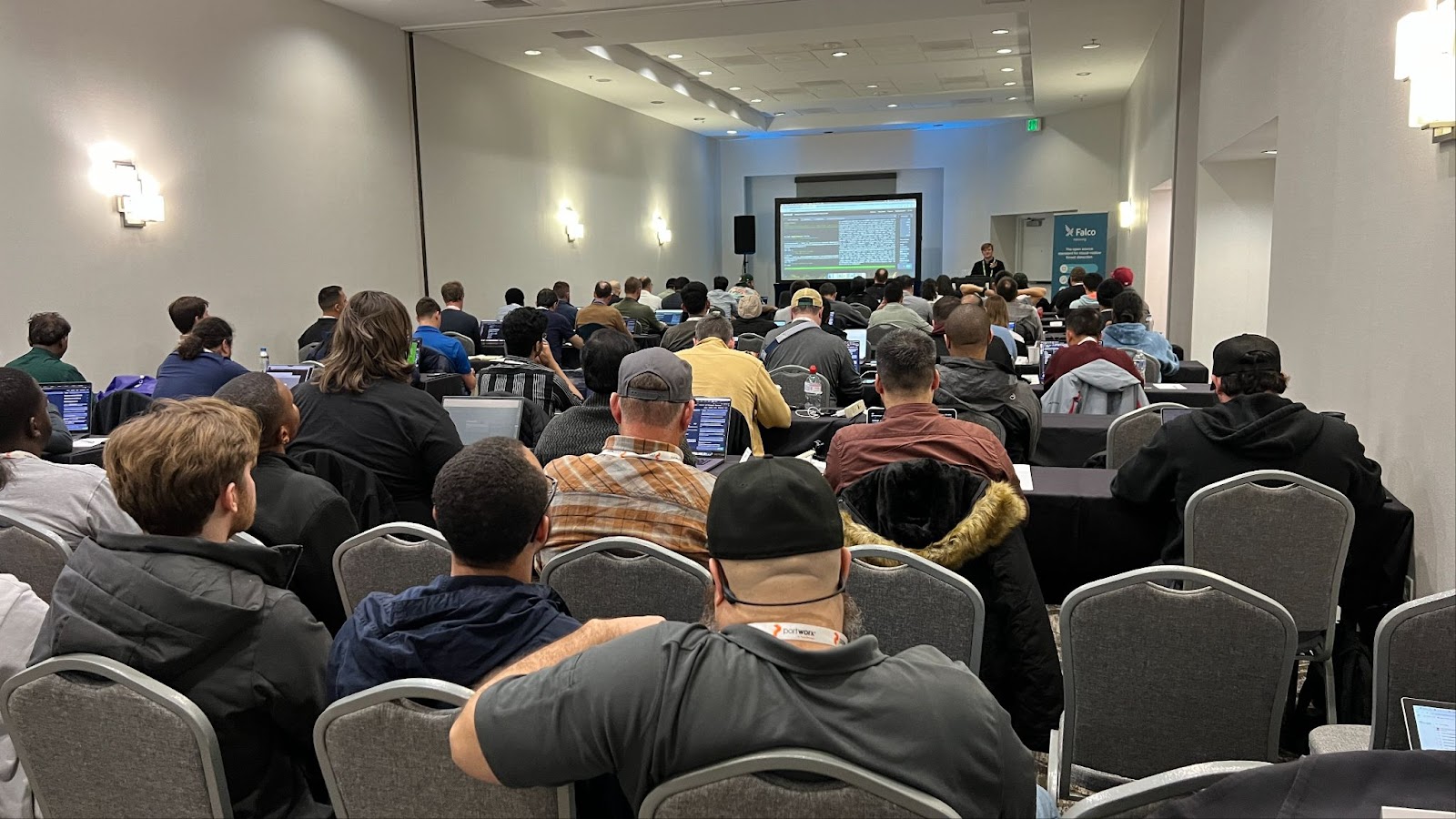A packed conference room attendees with laptops watch a Falco presentation on a projected screen.