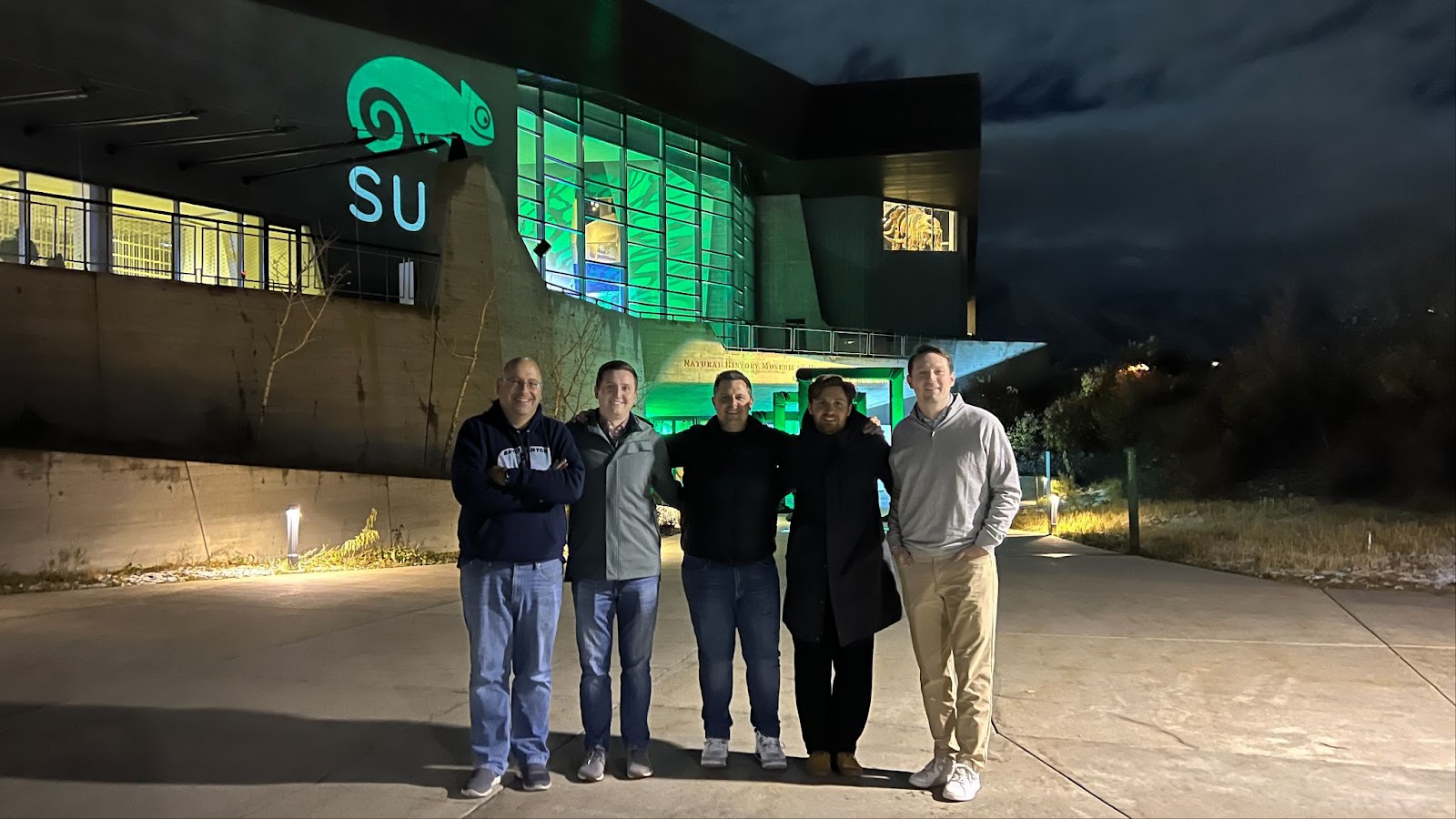 Five people pose together at night outside a Natural History Museum lit with an openSUSE chameleon logo.
