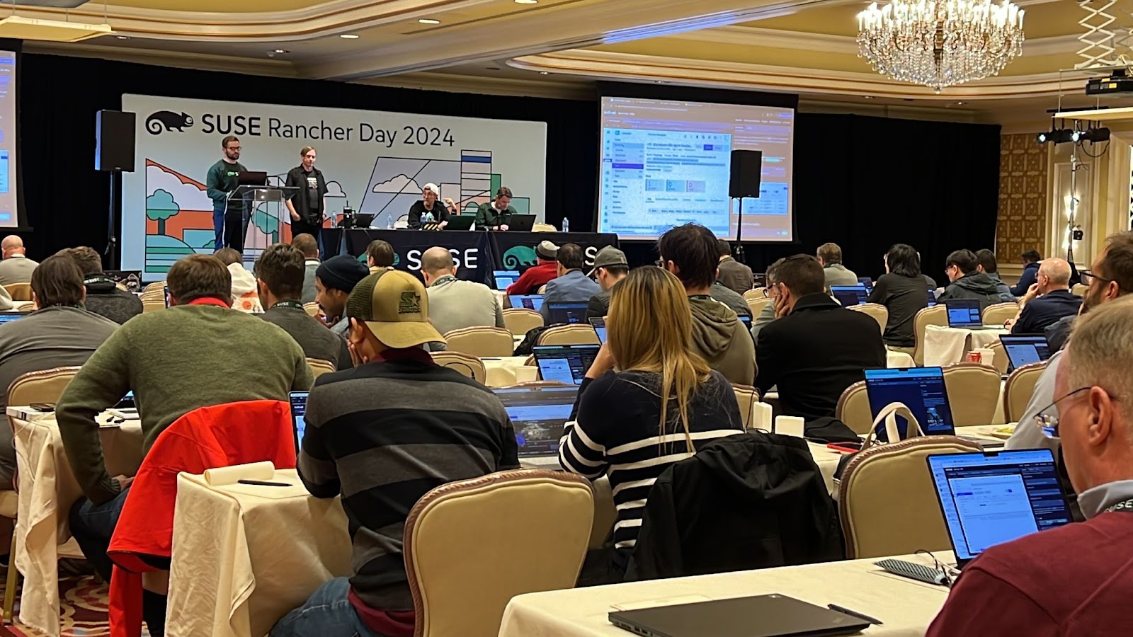 Attendees working on laptops at SUSE Rancher Day 2024, with presenters on stage at a ballroom venue.