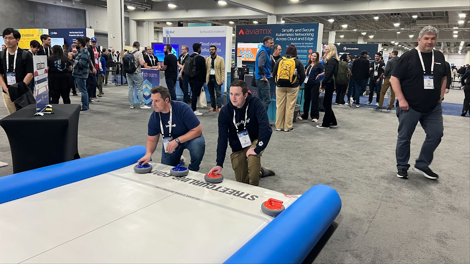 Two attendees play street curling on the expo floor at KubeCon, surrounded by vendor booths including Aviatrix and Buf.
