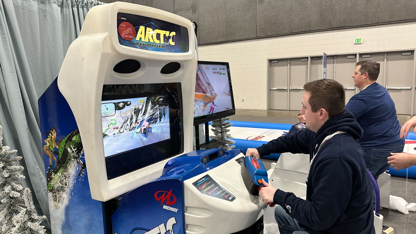 Two attendees playing Arctic Thunder, a Midway snowmobile racing arcade cabinet, at a convention hall.