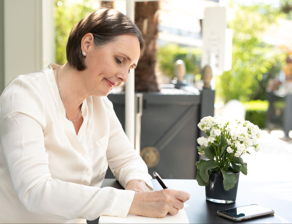 Woman in white blouse writing at an outdoor table with a potted white flower and smartphone.