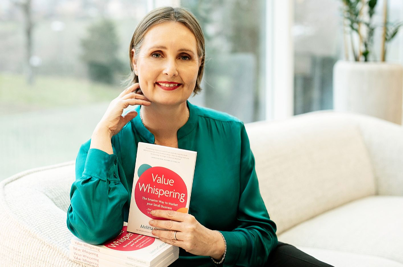 Woman in green blouse holding book titled 'Value Whispering' while sitting on a beige couch indoors.