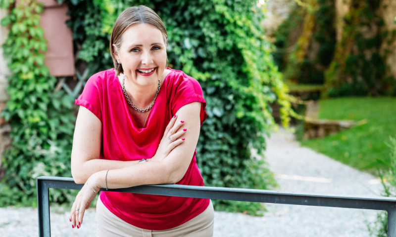 Smiling woman in a red blouse leaning on a metal railing with green ivy and garden in background.