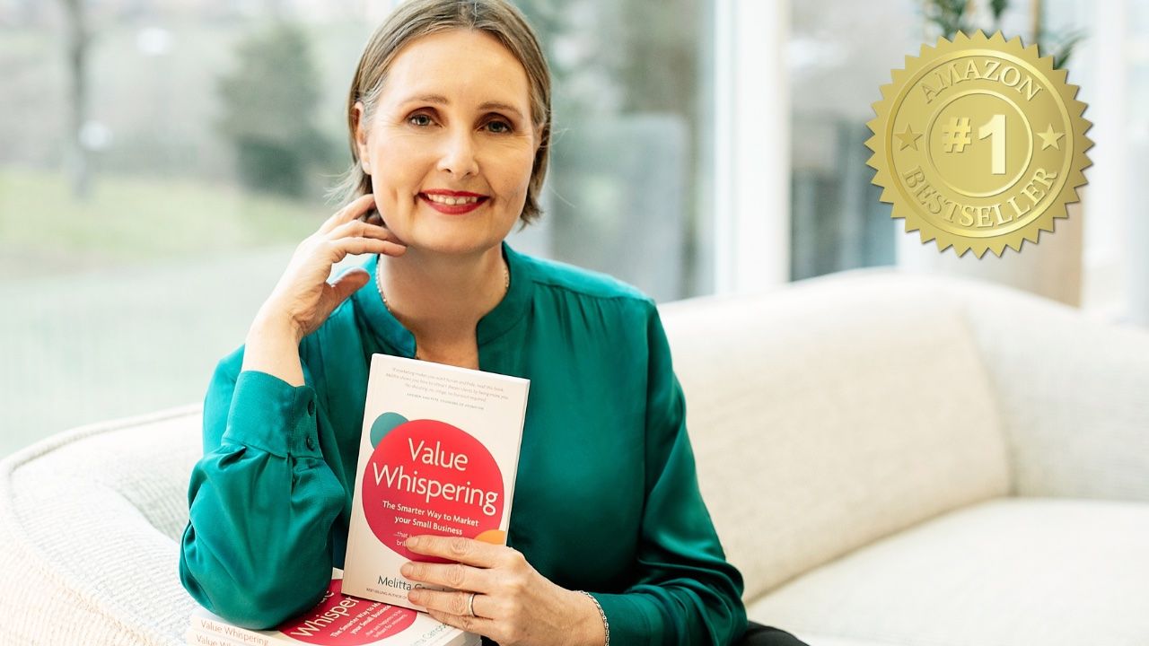 Woman in green blouse holding book titled 'Value Whispering' while sitting on a beige couch indoors.