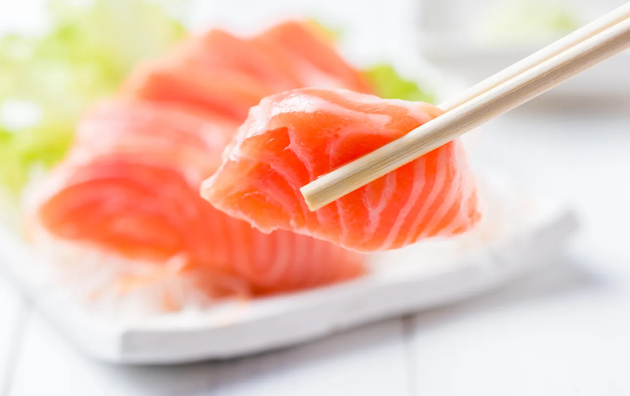 Ein besonders frisches Stück Lachs in Sashimi-Qualität, das elegant von zwei Stäbchen gehalten wird und mit seiner leuchtenden Farbe und zarten Textur überzeugt.