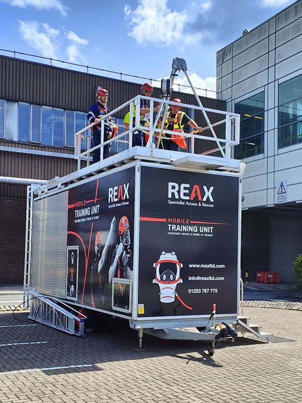 A black mobile training unit trailer labeled 'REAX Specialist Access & Rescue.' Three people wearing hard hats and safety harnesses are standing on a railed platform on the roof of the trailer, operating a tripod and winch system. The side of the trailer features graphics of a respirator mask and safety equipment, along with the text 'Mobile Training Unit - Confined Space & Work at Height' and contact information.