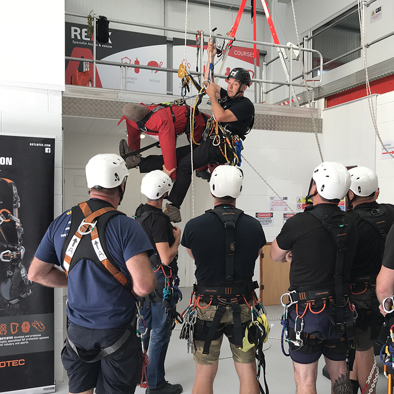 An indoor rescue training session showing an instructor suspended by ropes, demonstrating a mid-air rescue maneuver with a red training dummy. Below, a group of five trainees in white helmets and safety harnesses watch the demonstration.