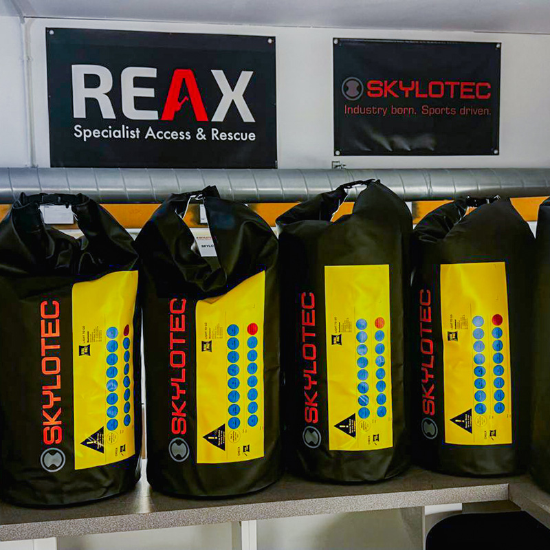 Four large, black Skylotec waterproof gear bags with yellow informational panels standing in a row on a counter. Above them hang black banners displaying the REAX and Skylotec logos.