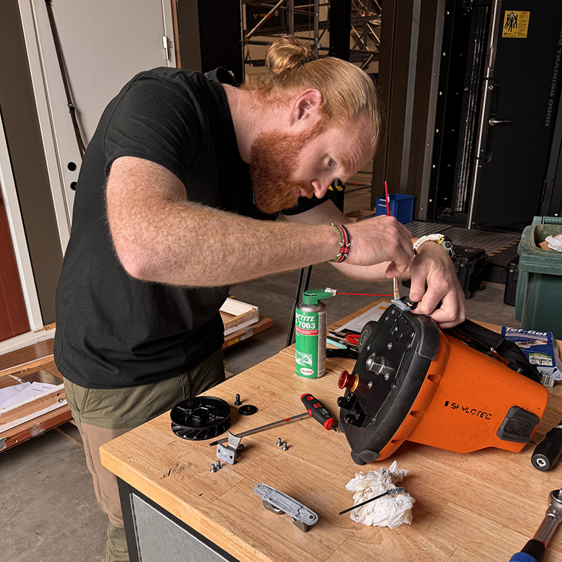 A man with a red beard works at a wooden workbench, carefully servicing the opened casing of an orange Skylotec power ascender using a small brush. Various hand tools, a can of Loctite, and loose parts surround the device on the bench.