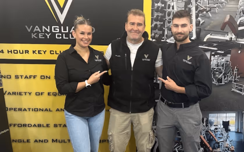 Three Vanguard Key Clubs team members standing in front of a branded gym display, smiling and pointing toward the center.