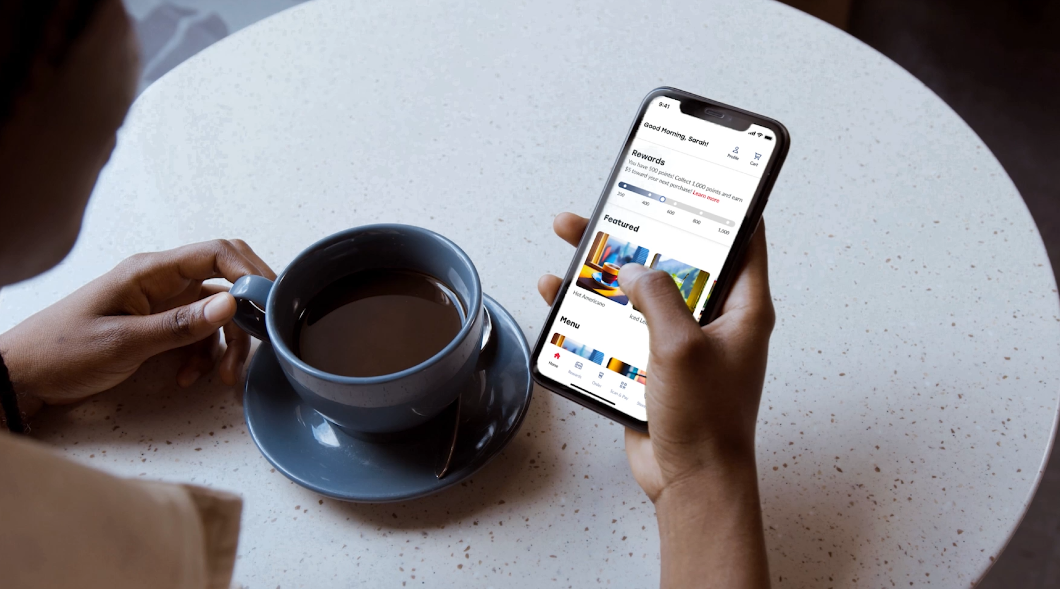 Overhead view of a person's hands, one holding a cup of coffee and the other browsing on a smartphone. The phone's screen displays a user interface with a greeting message 'Good Morning, Sarah,' followed by a rewards section and colorful, graphically rich feature banners below. The coffee cup is on a saucer, and both are placed on a speckled white table. The scene suggests a relaxed morning routine with technology and a warm beverage.