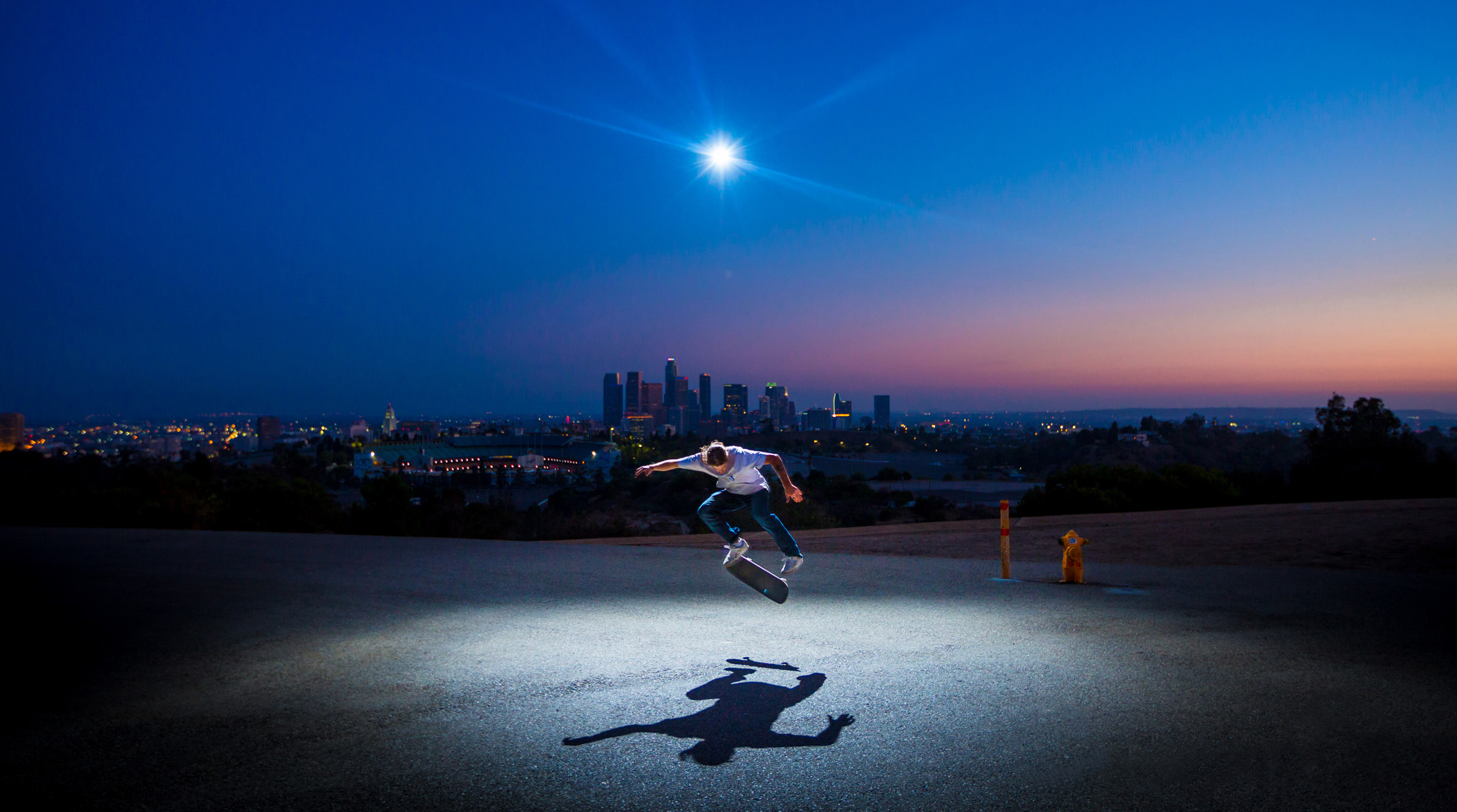 Skateboarder performing a trick at dusk with city skyline and bright light overhead.