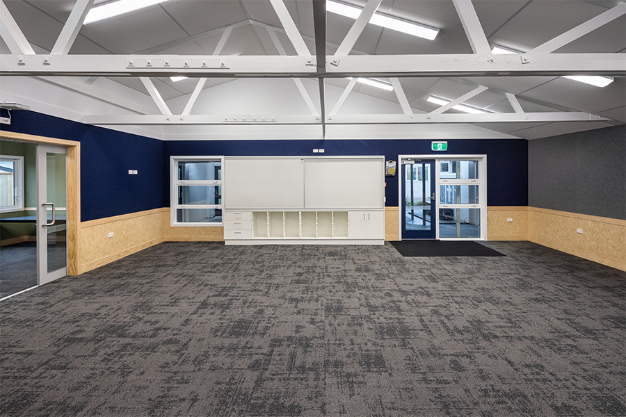 Modern classroom interior with exposed white trusses, blue feature wall, carpeted flooring, and built-in timber joinery