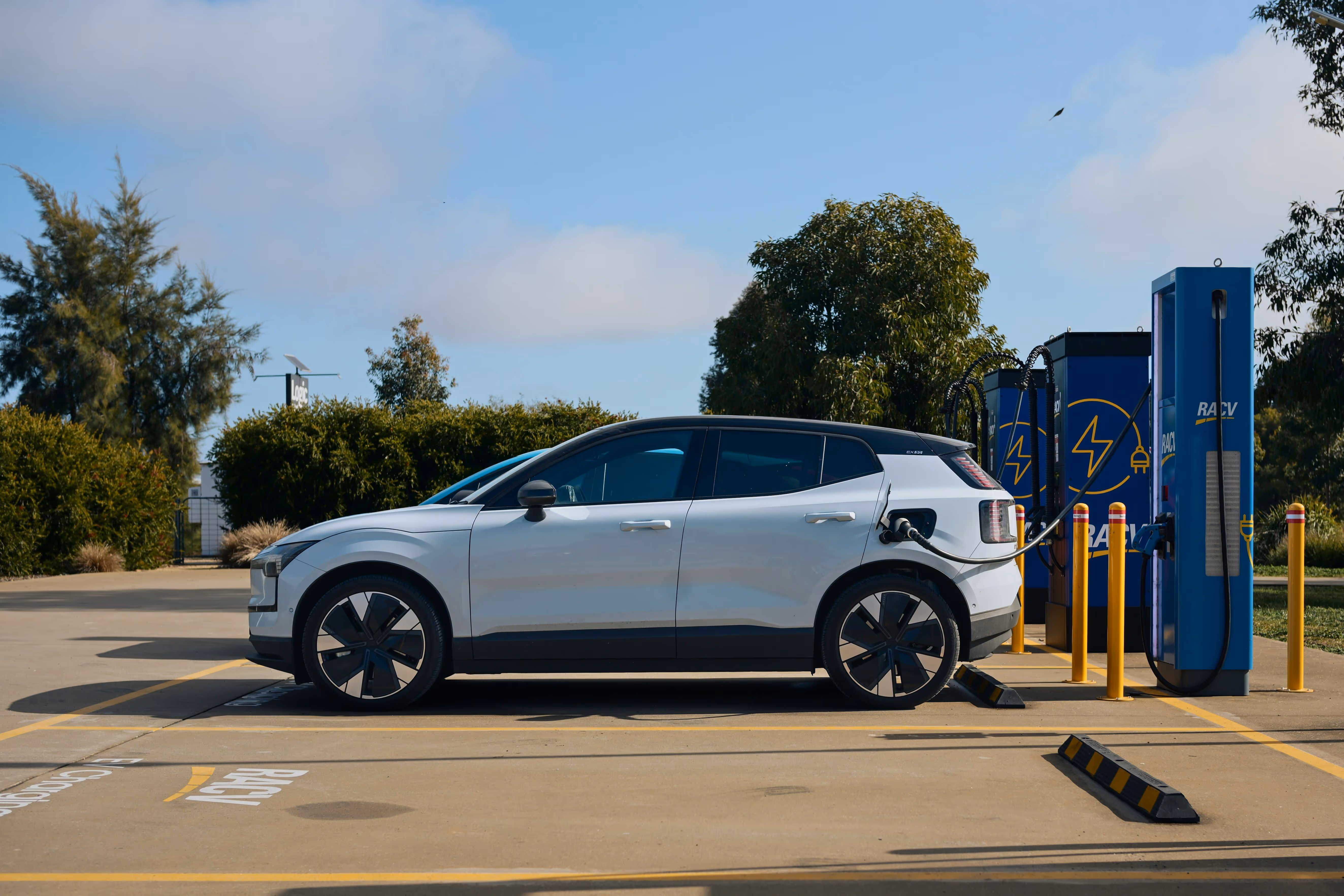 White electric car charging at a blue RACV charging station in a parking lot.