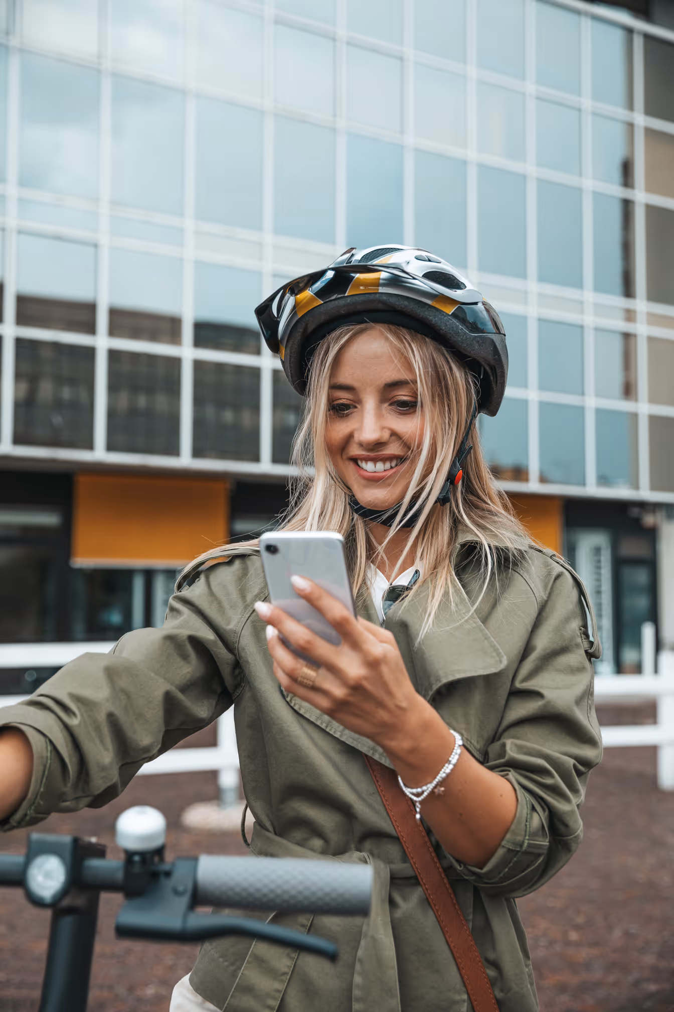 Smiling woman wearing a bike helmet and green jacket looking at her phone while holding an electric scooter handle.