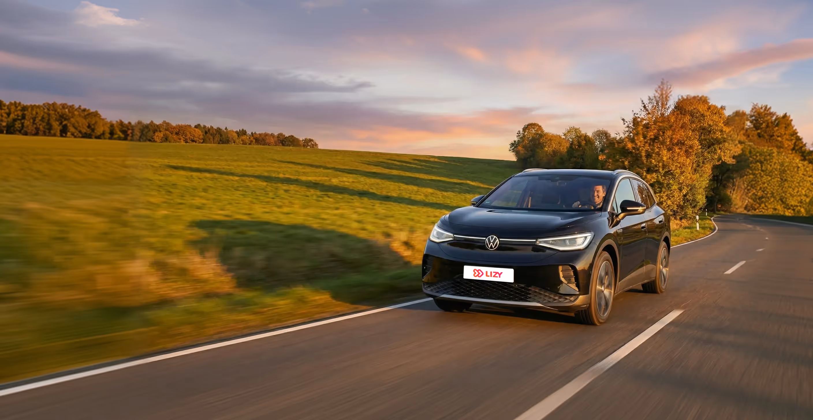 Black Volkswagen SUV driving on a countryside road at sunset with a smiling driver.