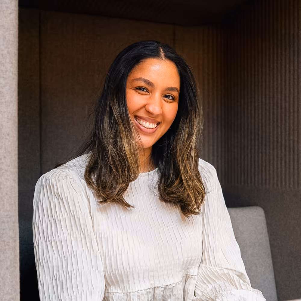 Smiling woman with long dark hair wearing a white textured blouse sitting against a dark background.