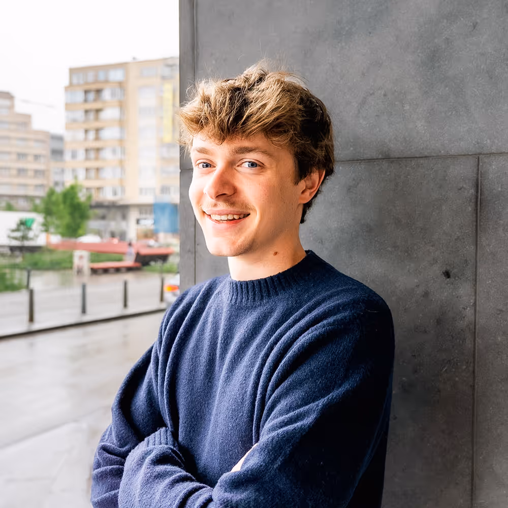 Jeune homme souriant aux cheveux châtain clair, portant un pull bleu marine, debout devant un mur en extérieur.