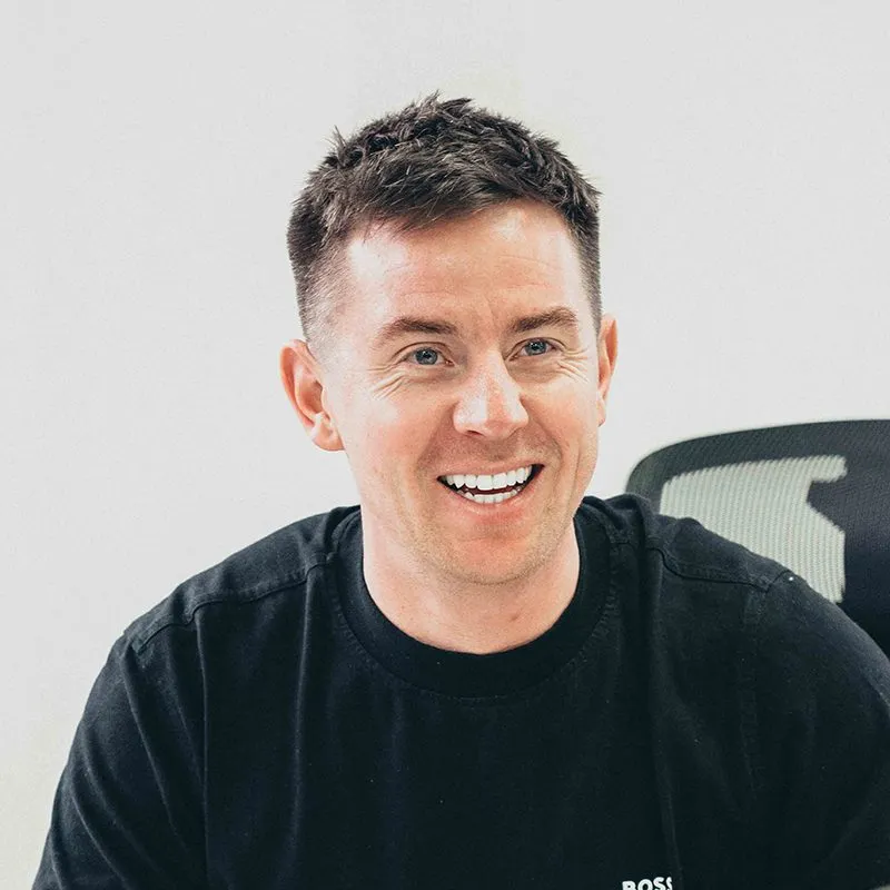 Smiling man with short brown hair wearing a black Boss T-shirt, seated in front of a plain light background.