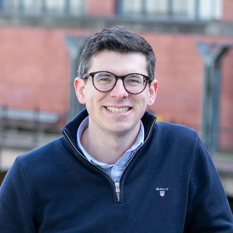 Smiling man with dark hair and glasses wearing a navy blue quarter-zip sweater outdoors.