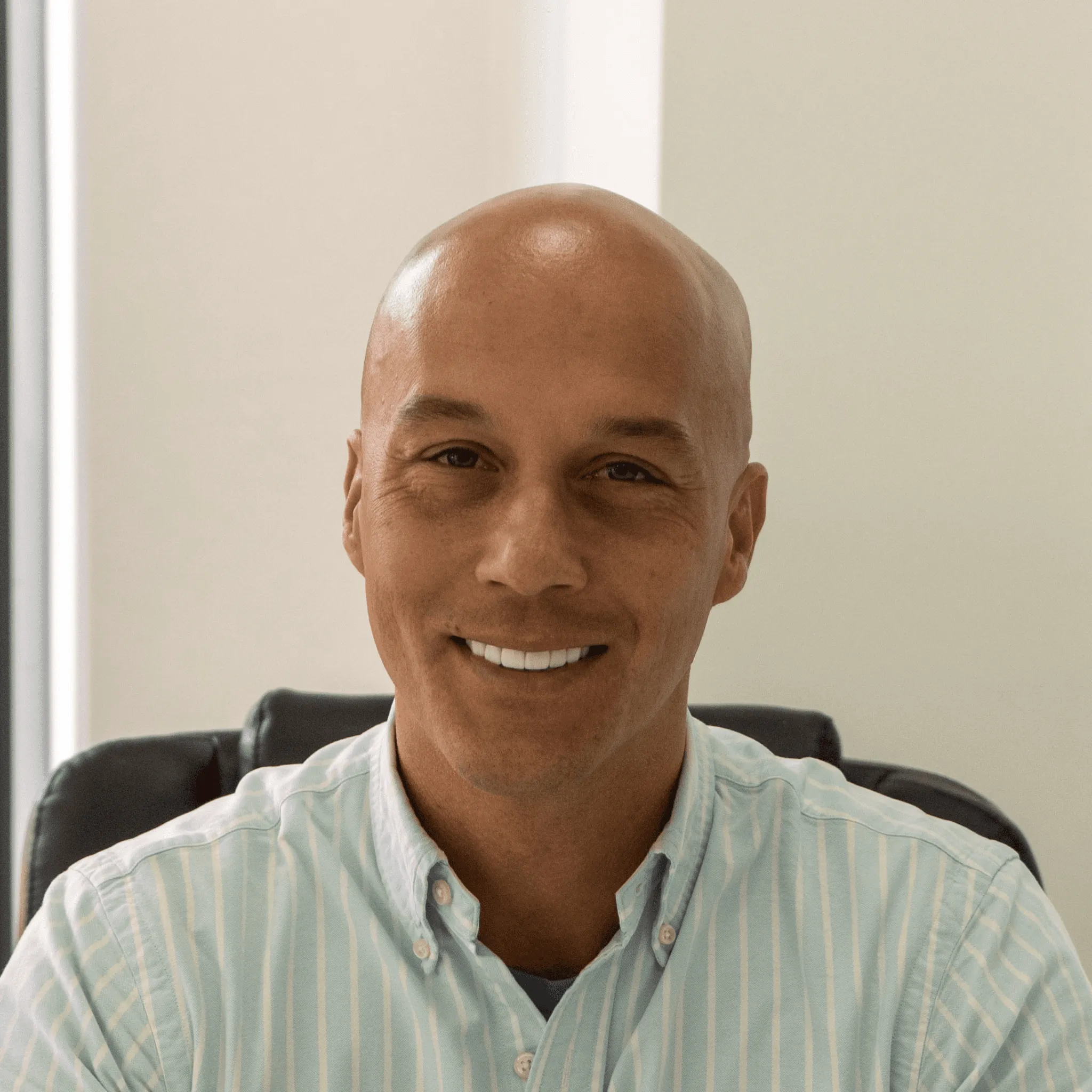Smiling bald man wearing a light blue striped button-up shirt seated indoors.