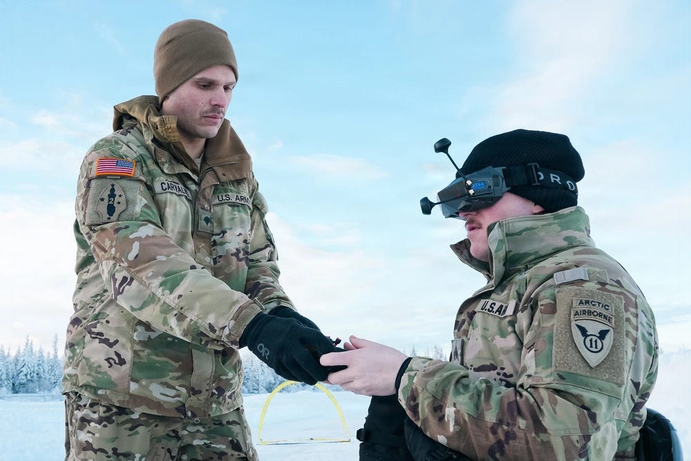Two U.S. Army soldiers are engaged in a drone operation exercise in a snowy, arctic environment. The soldier on the right is wearing FPV (First-Person View) drone goggles, while the soldier on the left, identified by name tape as 'CARTAL' and wearing a 2nd Infantry Division patch, appears to be handing over a small drone or component. The setting and unit patches suggest this is a cold-weather training exercise.