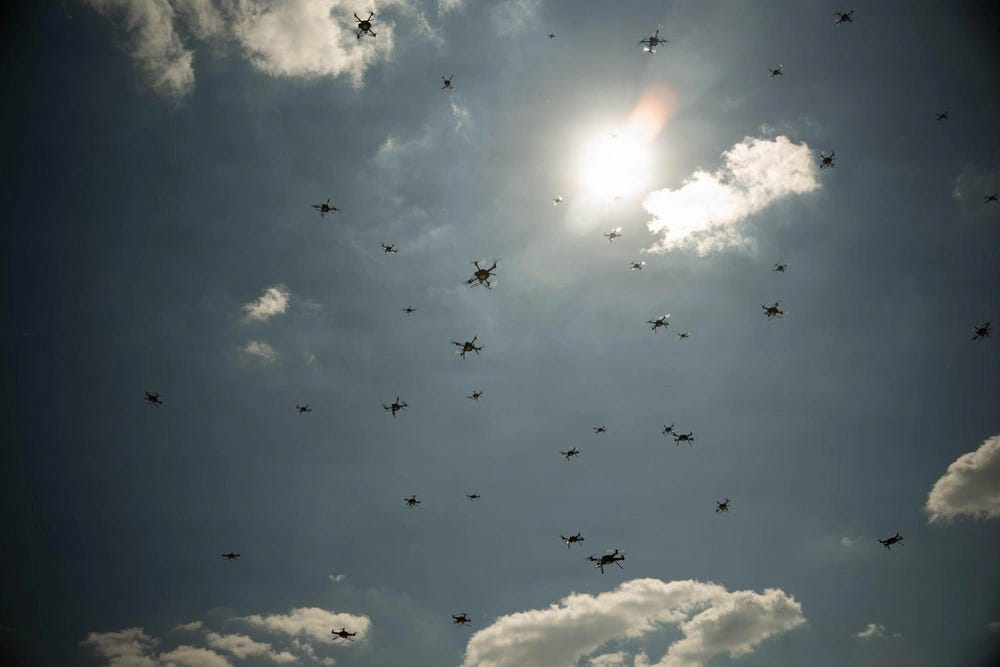 The image shows a large swarm of multirotor drones (quadcopters/hexacopters) filling the sky, photographed from a low angle looking upward. The drones appear as dark silhouettes against a partly cloudy sky with a bright sun visible near the center-upper portion of the frame. This appears to be a coordinated drone swarm demonstration or display event.