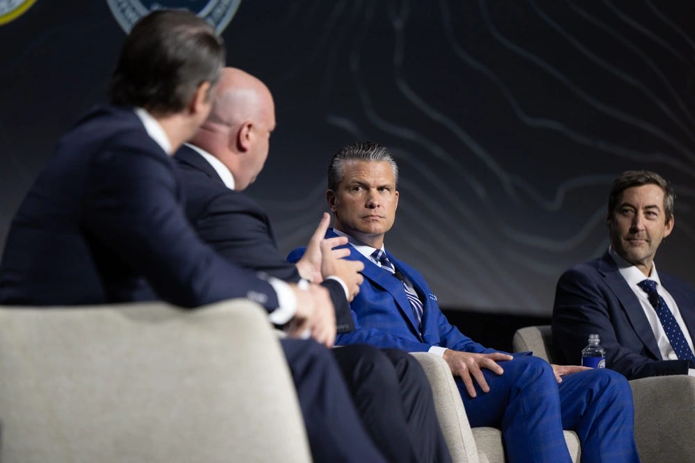 Four men in business suits are seated in chairs participating in what appears to be a formal panel discussion or conference. One man in a distinctive royal blue suit is prominently featured in the center, attentively listening to a speaker on the left. The setting includes a dark backdrop with what appears to be an official seal or logo.