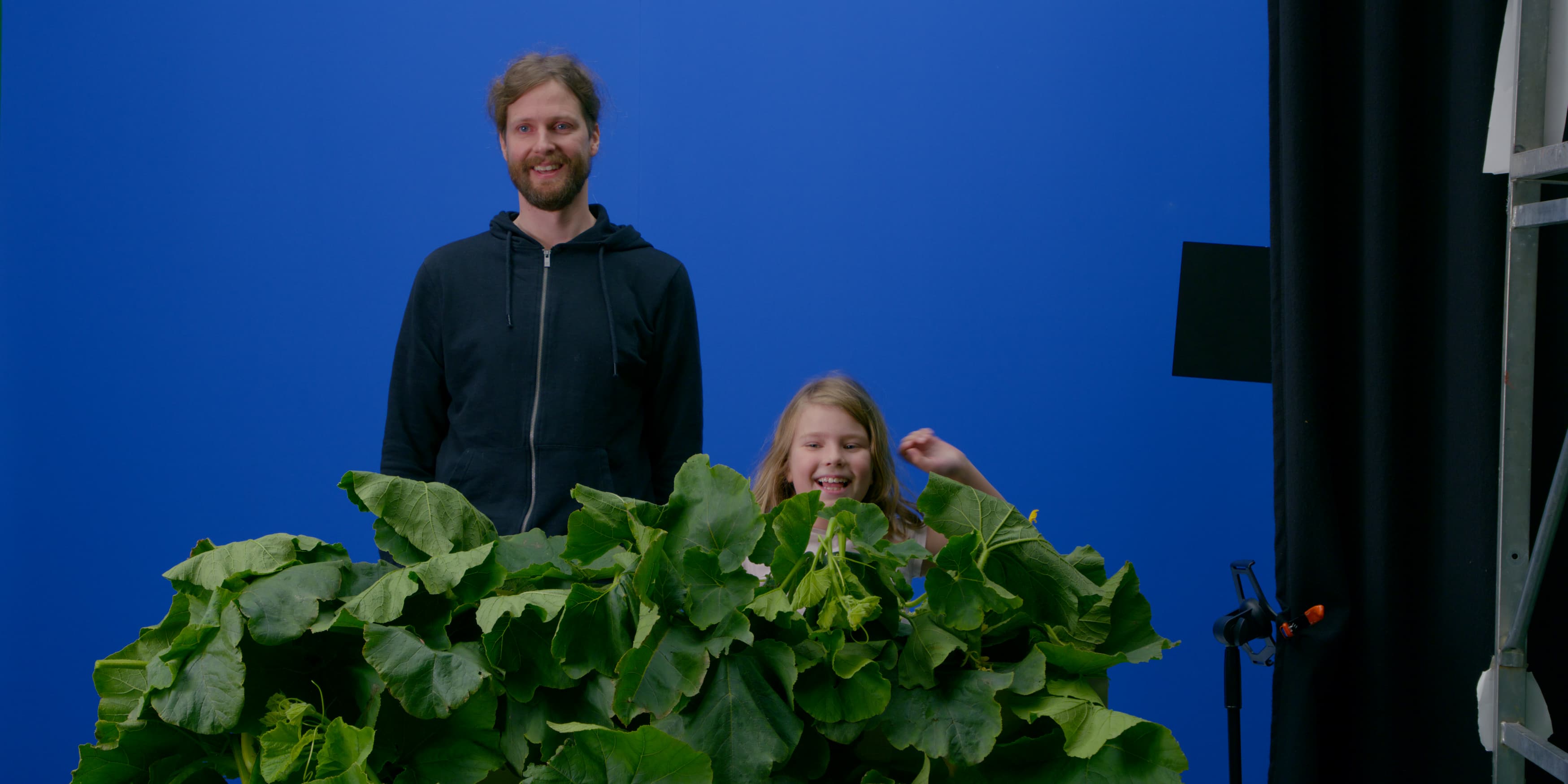 Två personer bakom stora gröna blad framför en blå bakgrund i studion