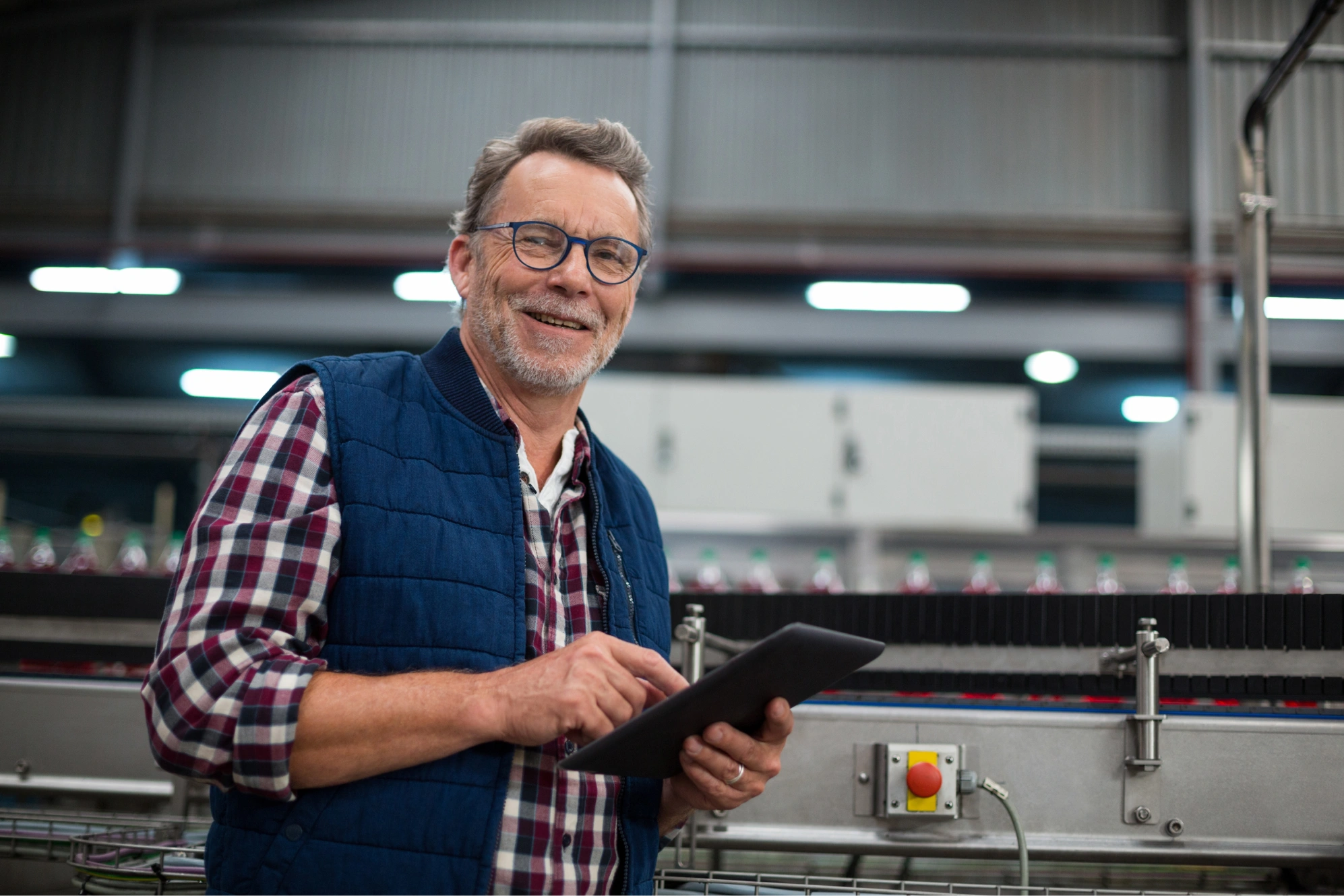 Smiling man wearing glasses and a blue vest using a tablet in an industrial factory setting.