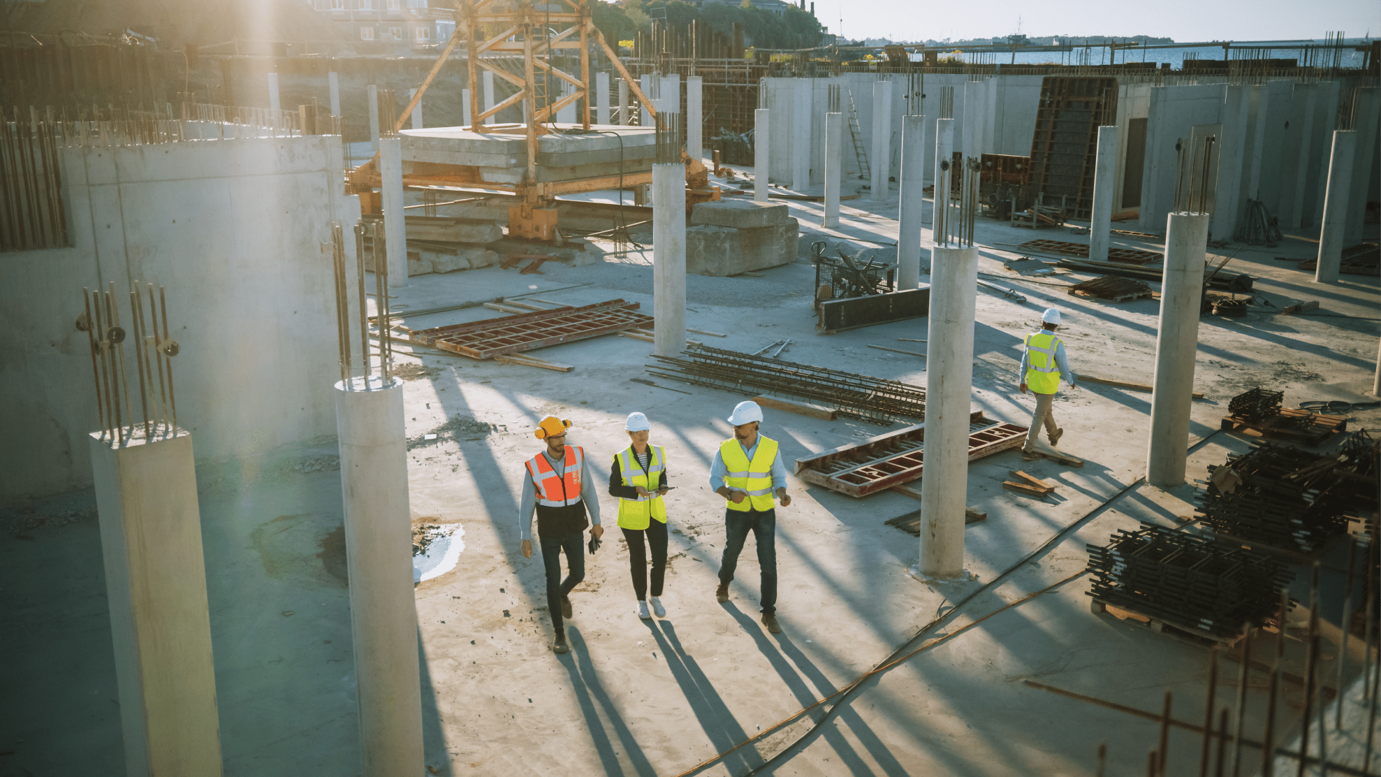 Four construction workers wearing hard hats and high-visibility vests at a building site with concrete pillars and construction materials, walking and inspecting the area in daylight.