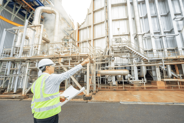 Engineer in a white helmet and neon yellow safety vest holding rolled blueprints and pointing at an industrial plant with large metal pipes and structures.