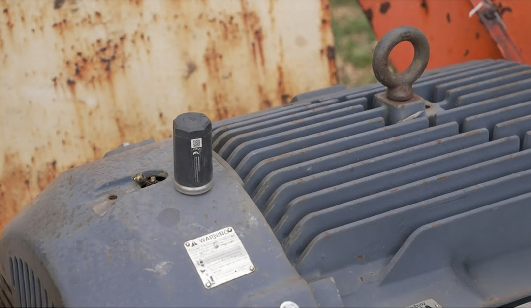 Close-up of a gray industrial machine with cooling fins and a cylindrical sensor attached on the top left side, featuring a QR code. The background shows a rusty surface and green grass.