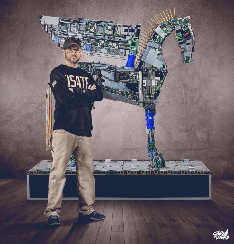 Man in a USATE sweatshirt standing with arms crossed in front of a horse sculpture made of computer parts