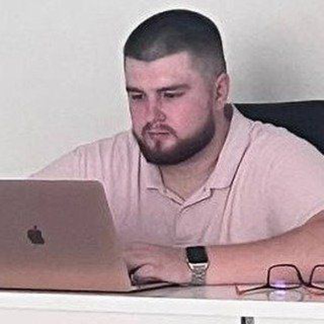 Bearded man in a beige polo typing on a MacBook, glasses on the desk