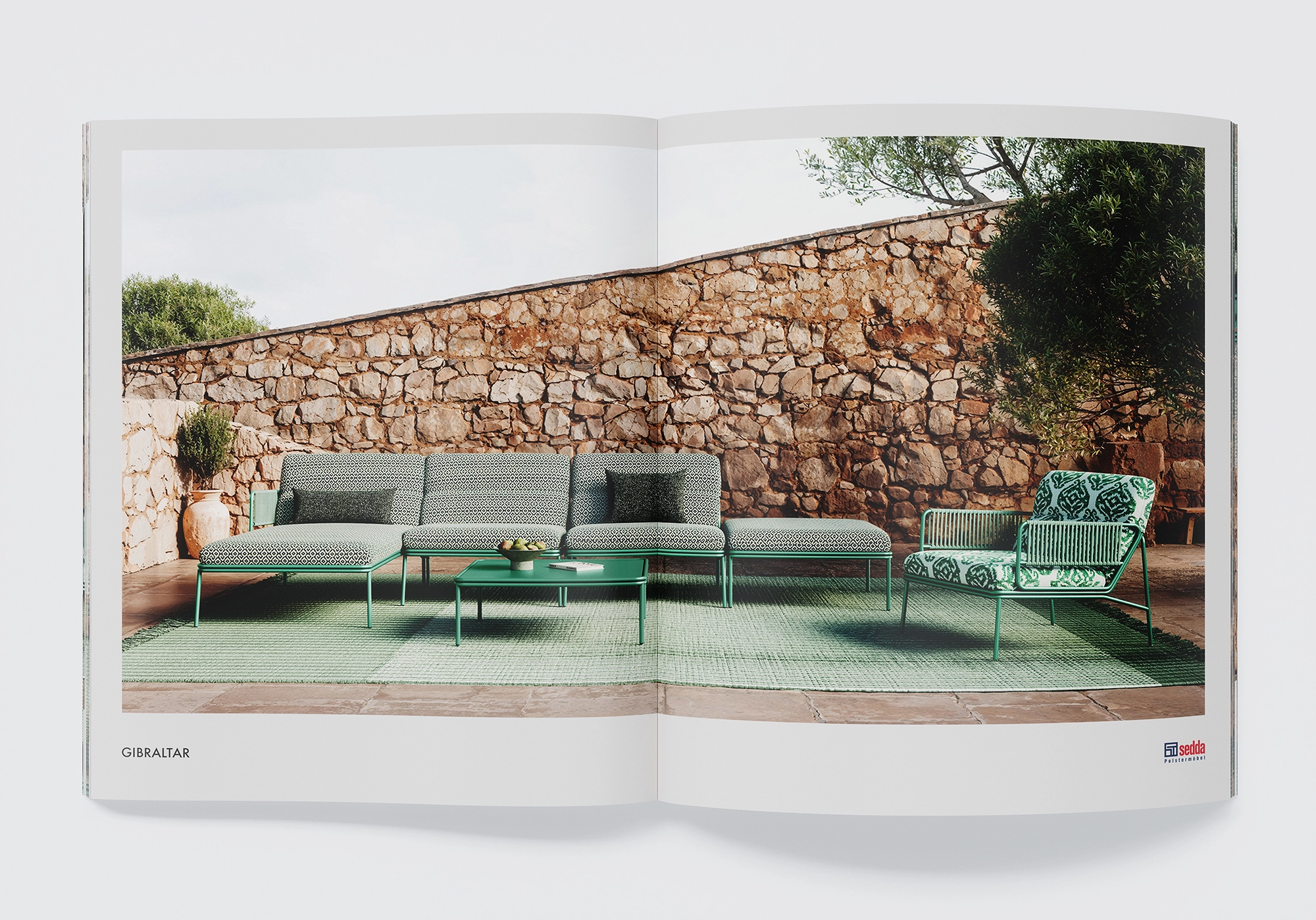 Visualizzazione 3D per mobili da giardino e prodotti per esterni: sedda "Gibraltar" con cuscini fantasia davanti a un muro di pietra - scena di patio mediterraneo.