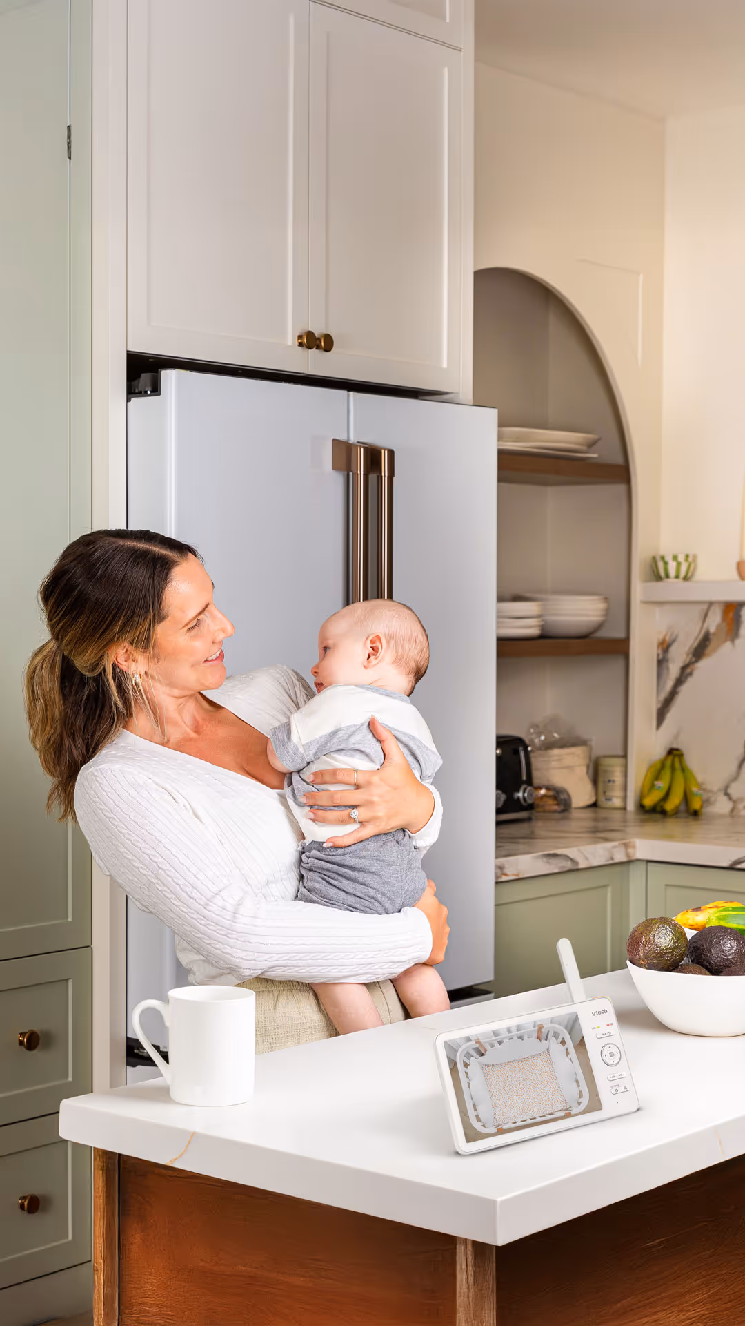 Woman holding and smiling at a baby in a modern kitchen near a white countertop with a baby monitor, mug, and bowl of avocados and bananas.