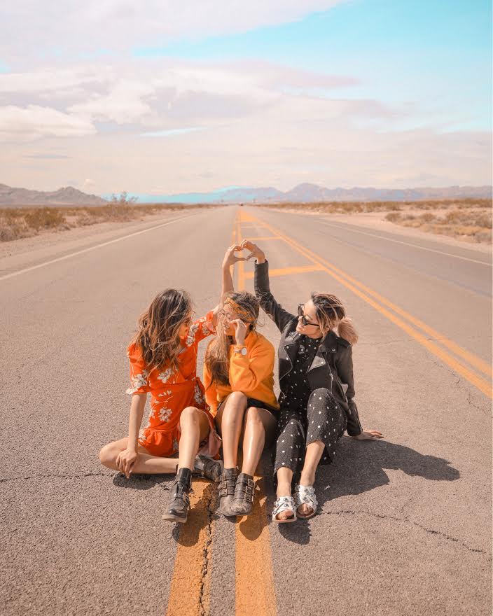 Three women sitting on a desert road making a heart shape with their hands under a partly cloudy sky.