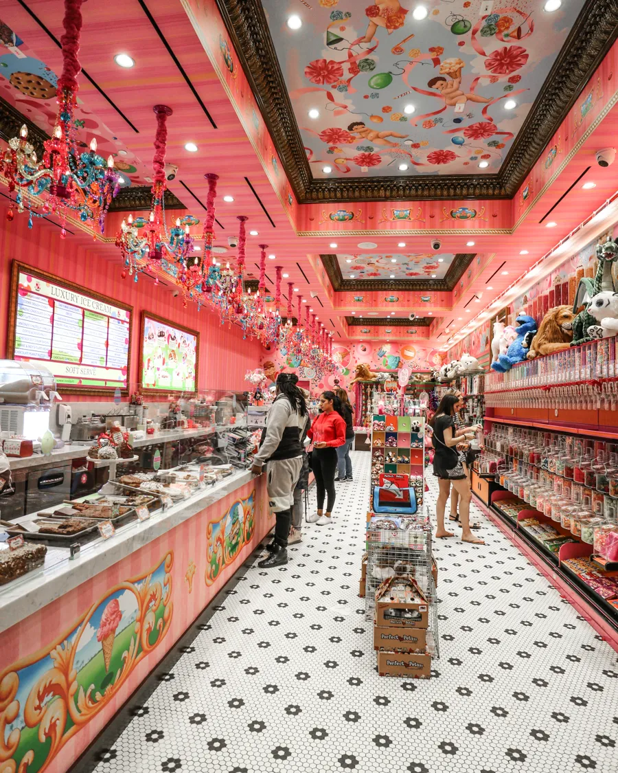 Colorful candy and dessert shop interior with pink walls, ornate ceiling paintings, and customers browsing sweets and treats.