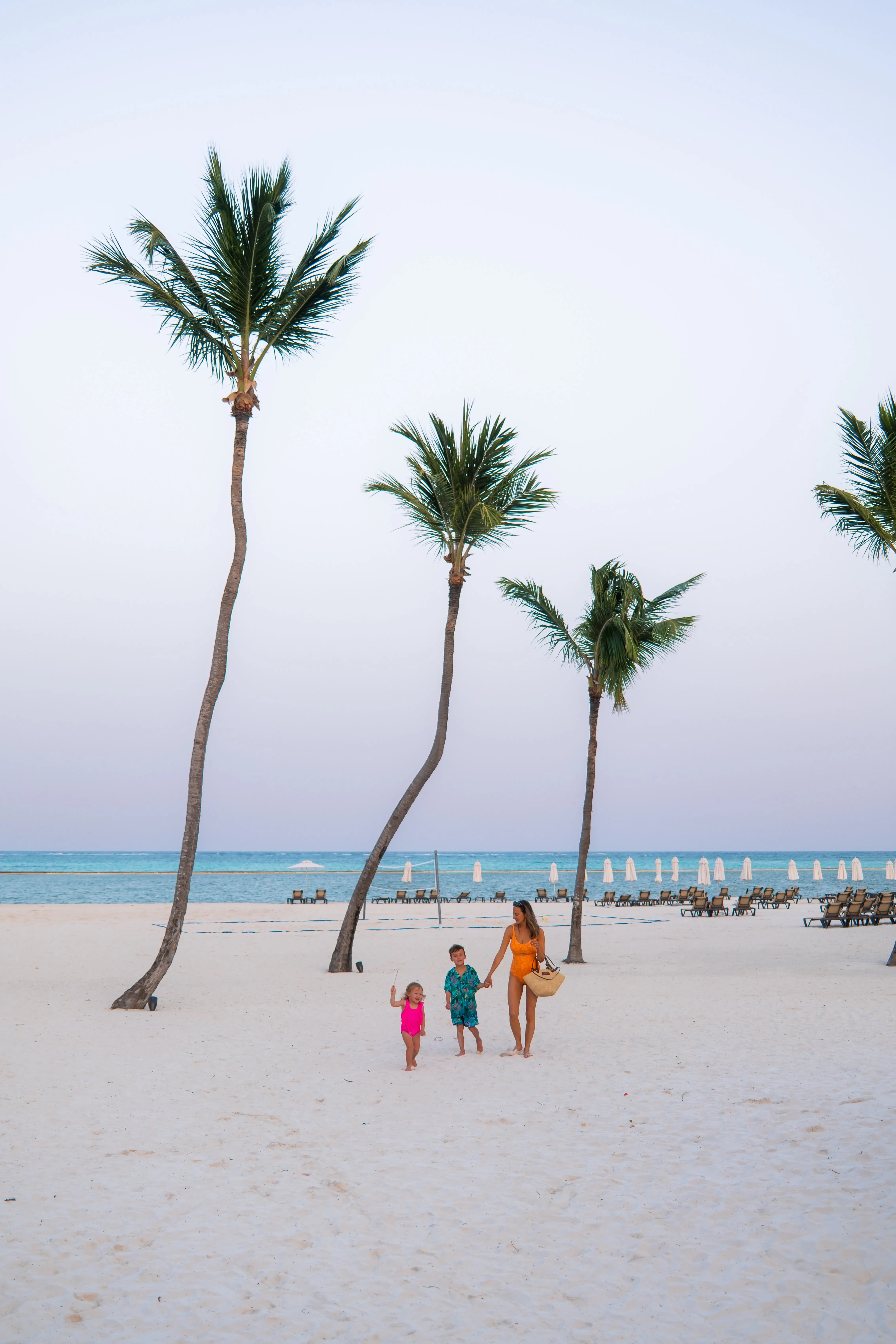 Woman in an orange swimsuit holding hands with two children walking on a sandy beach with palm trees and ocean in the background.