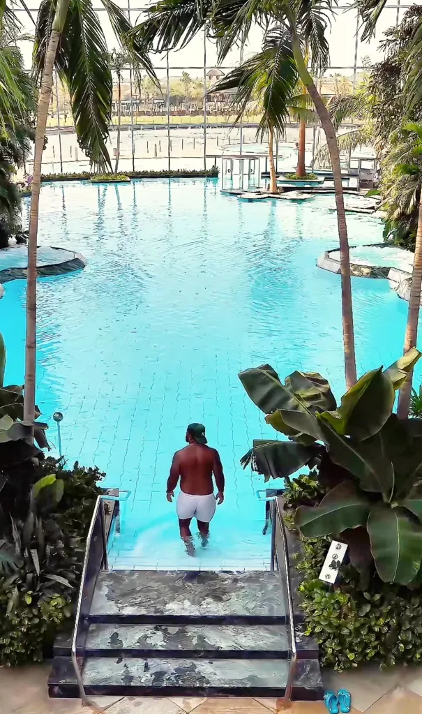 Man in white shorts and green cap entering a large indoor pool surrounded by palm trees and tropical plants.
