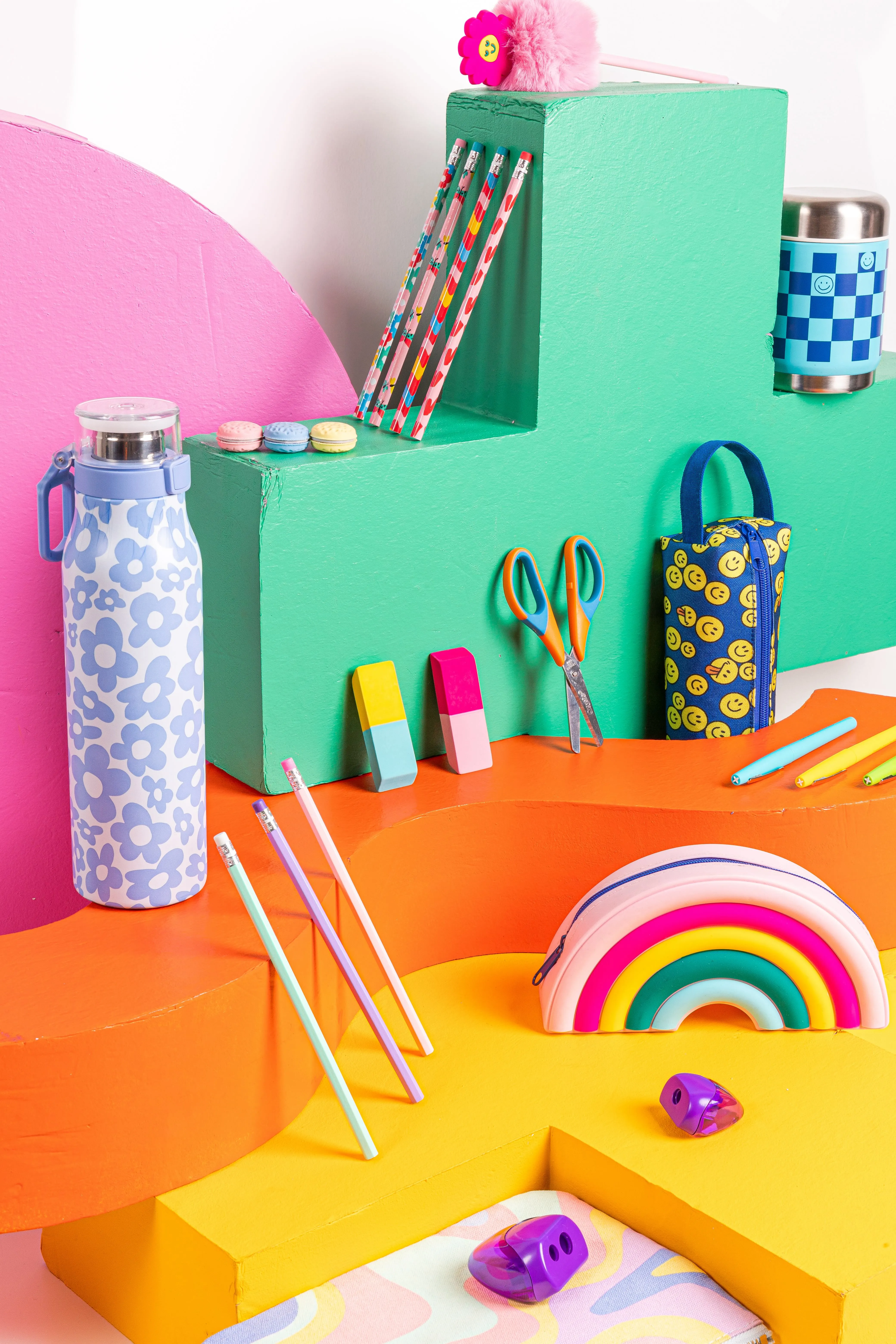 Colorful school supplies including patterned pencils, scissors, erasers, a rainbow pencil pouch, pencil sharpeners, and a water bottle displayed on green, orange, and yellow geometric blocks.