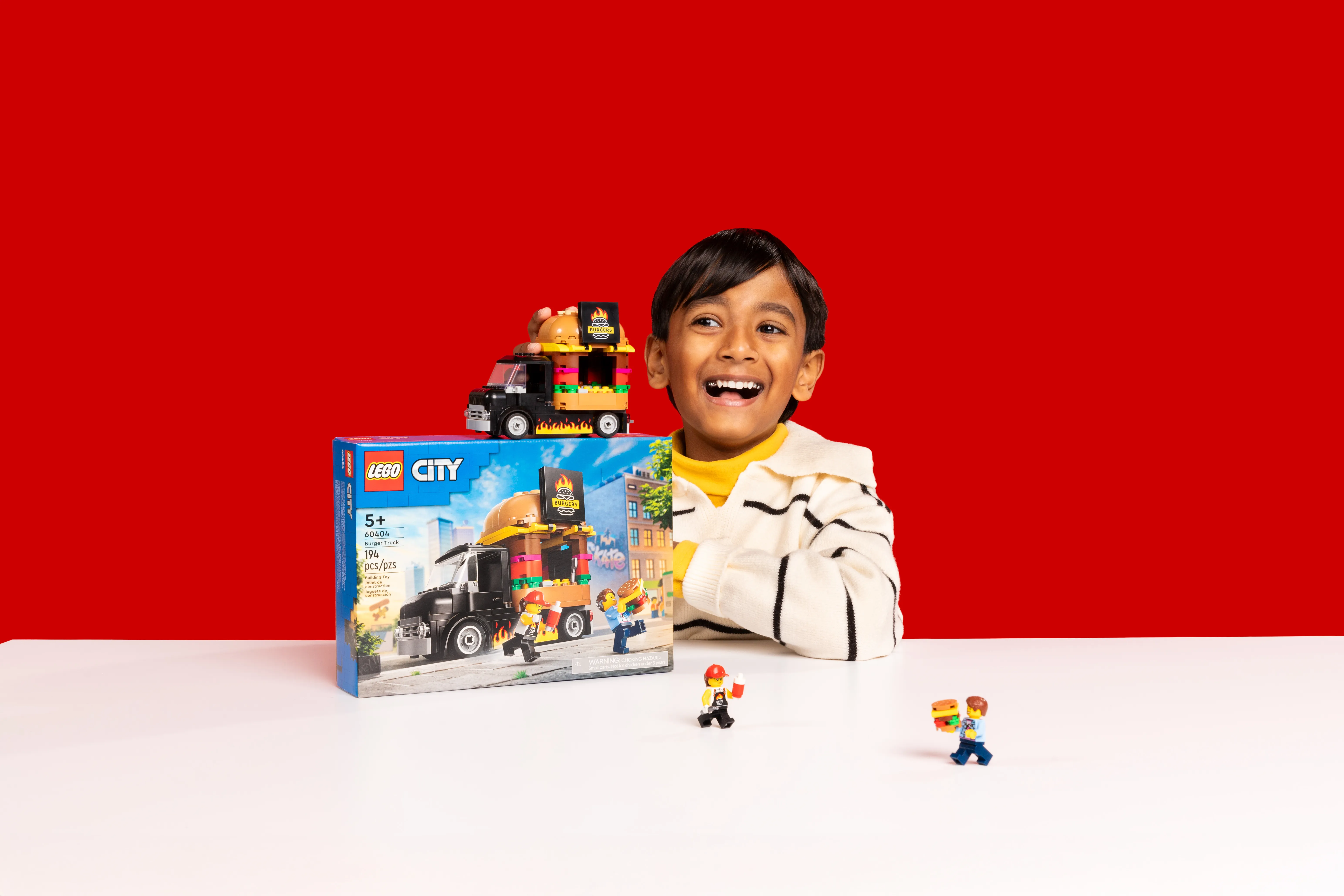Smiling boy playing with LEGO City Burger Truck set and minifigures on a white table against a red background.